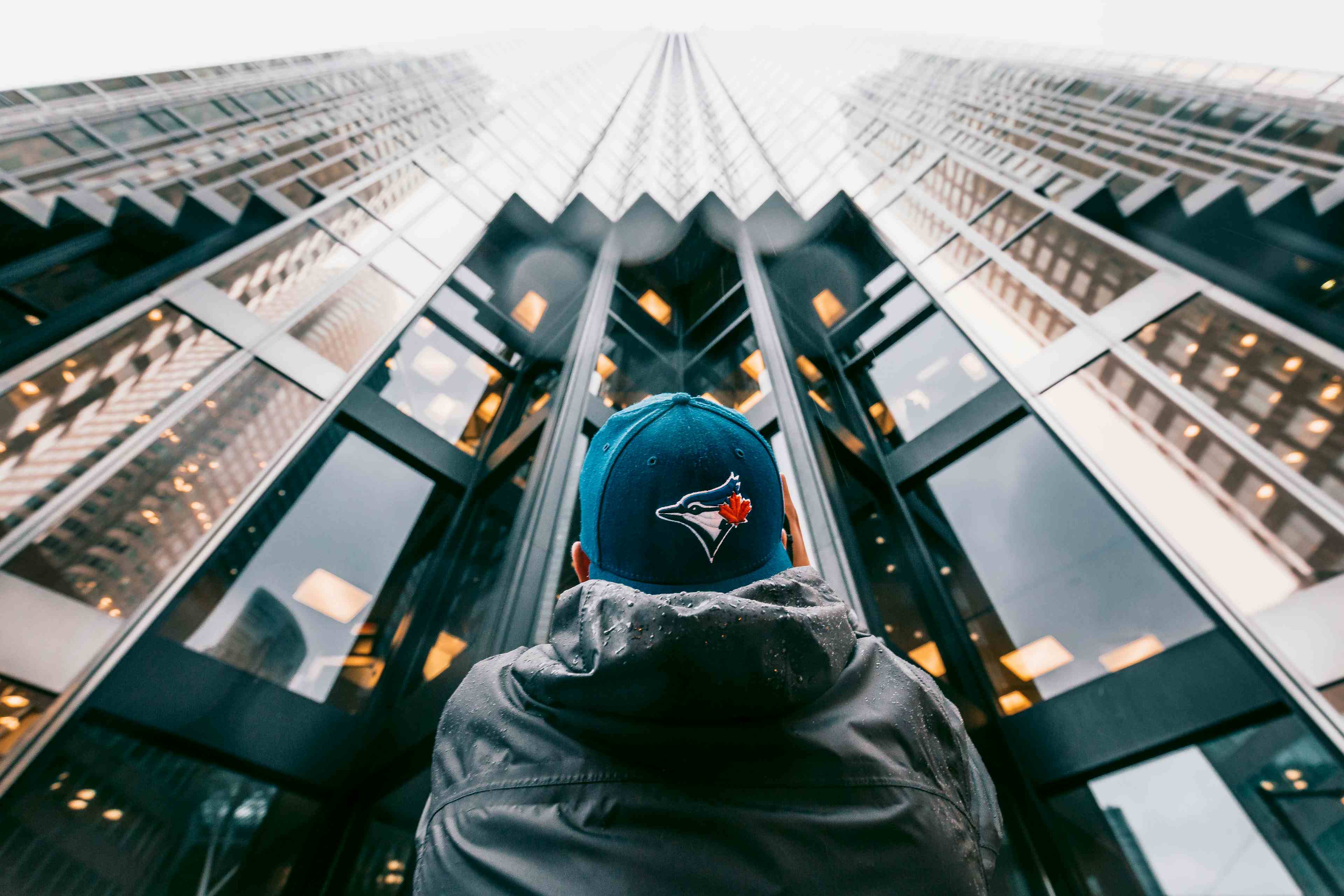 A man wearing a Blue Jays hat looking up at a golden coloured skyscraper in Toronto, Canada with a camera in his hand